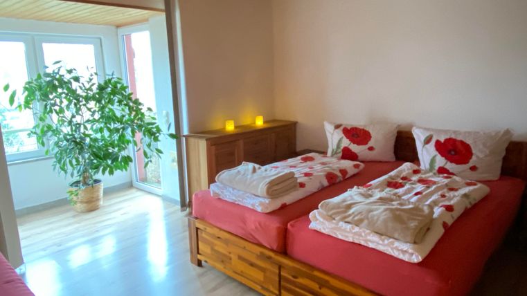 Ein helles Schlafzimmer mit zwei Betten, die mit roten Kissen und Decken dekoriert sind. Im Raum steht eine große Pflanze und es gibt eine angenehme, einladende Atmosphäre.
