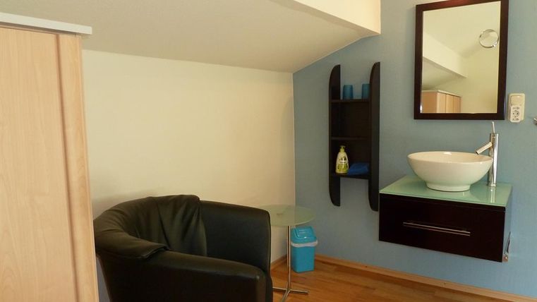 A small room with a black armchair and a sink. The walls are painted blue and there is a mirror as well as a shelf.