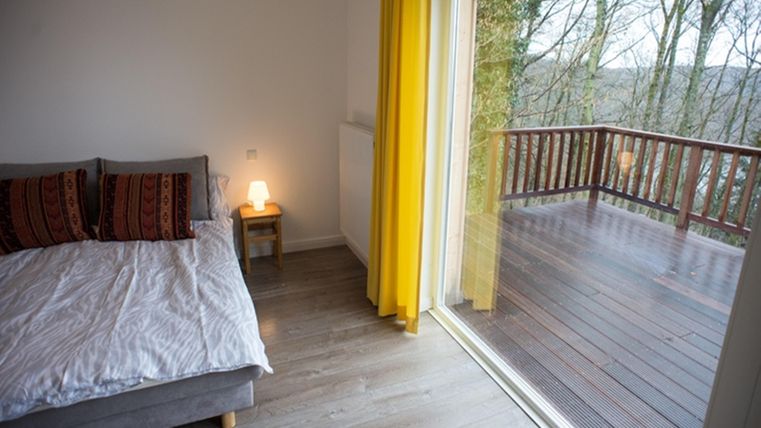 A cozy bedroom with a bed and colorful pillows. Large windows offer a view of the balcony and the trees outside.