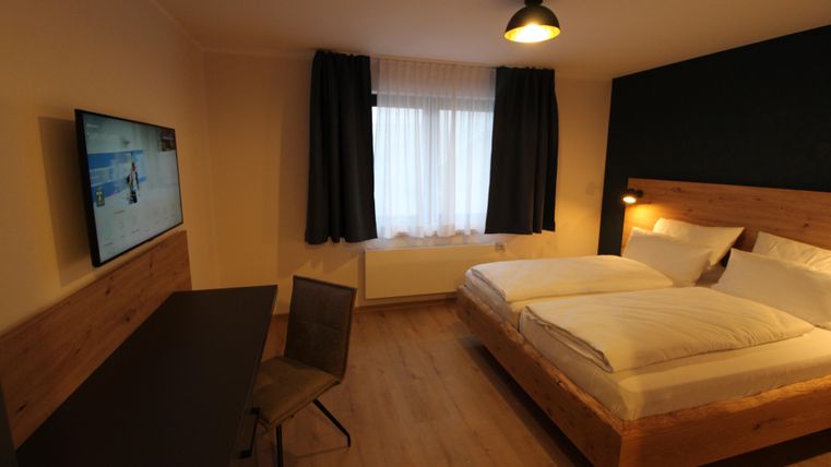 A modern bedroom with two beds and a desk. The room includes a television and large windows with curtains.