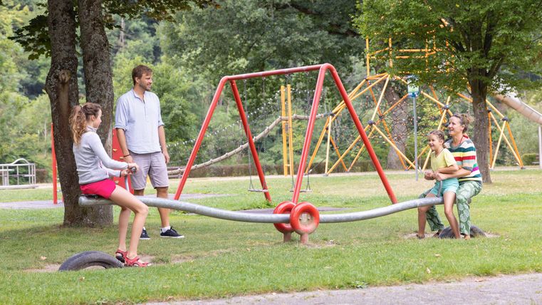 A playground with a seesaw and two groups of children. In the background, swings and a lot of green grass can be seen.
