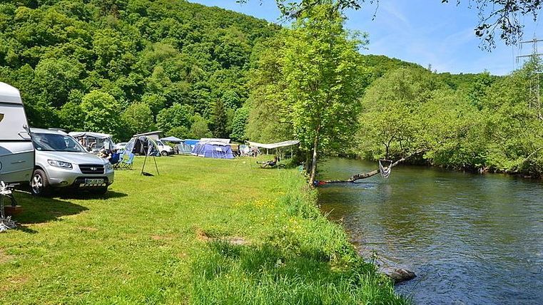 Ein grünes Campinggebiet neben einem ruhigen Fluss. Es gibt Wohnmobile und Zelte, umgeben von Bäumen und Wiese.