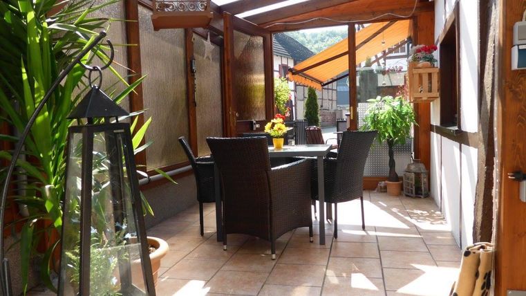 Een gezellige terrassenruimte met een tafel en stoelen van riet. De omgeving is groen met planten en een grote lantaarn.