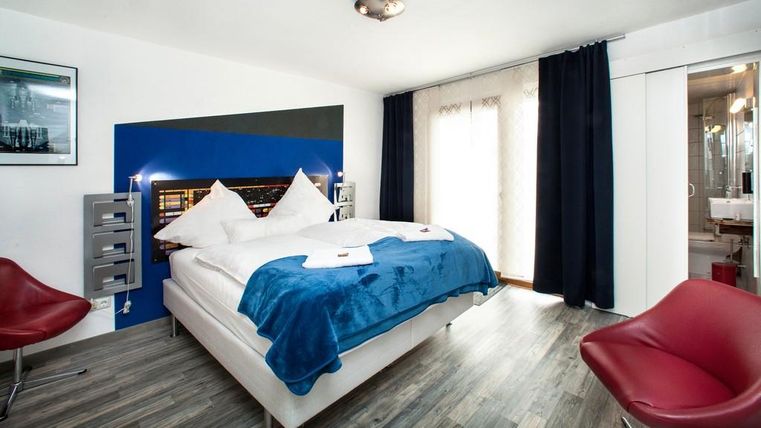 Ein modernes Schlafzimmer mit einem großen Bett und blauen Akzenten. Rote Stühle ergänzen die stilvolle Einrichtung und der Raum hat große Fenster für viel Licht.