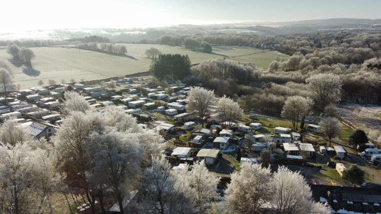 A campsite with many motorhomes and caravans is surrounded by frost-covered trees. In the background, there are wide meadows and a clear sky.