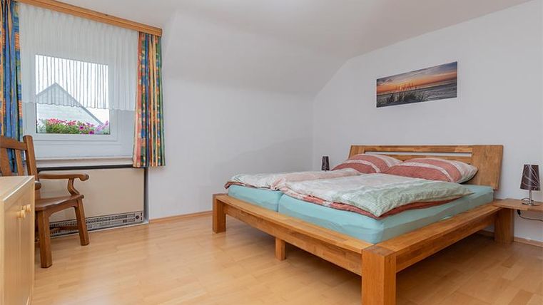 Ein helles, gemütliches Schlafzimmer mit einem Holzbett und bunten Vorhängen. Neben dem Bett steht ein Stuhl und ein kleines Bild hängt an der Wand.