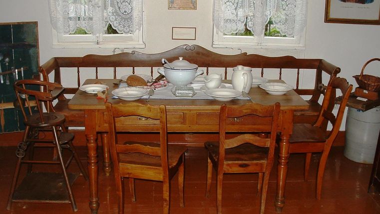 A nostalgic dining area with a large wooden table and six chairs. On the table are dishes and a soup plate, surrounded by bright windows with curtains.