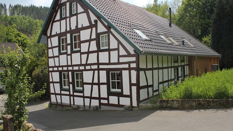 A lovely half-timbered house with a sloping roof stands on a quiet street. Surrounded by green trees and grass, it appears inviting and traditional.