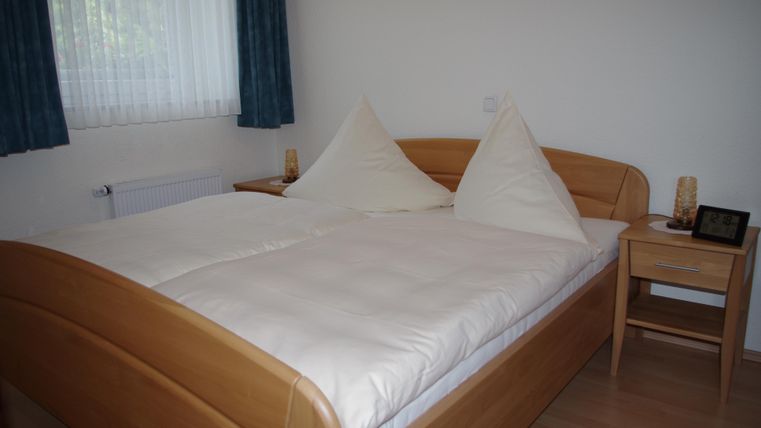 A cozy bedroom with a wooden double bed and beautiful bedding. Next to the bed is a bedside table with a lamp and a small screensaver.