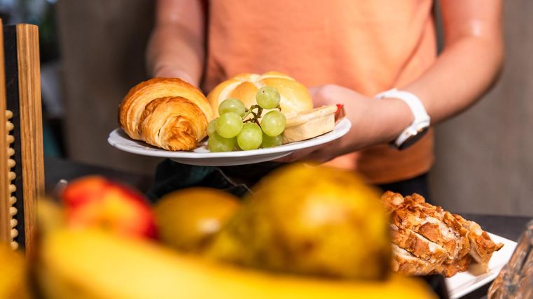A person is holding a plate with fresh rolls, grapes, and a croissant. In the background, various fruits are visible.