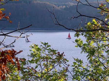 Een rustige zee, omringd door bomen, met twee zeilboten op het water. De kleurrijke bladeren op de voorgrond geven de scène een herfstachtige sfeer.