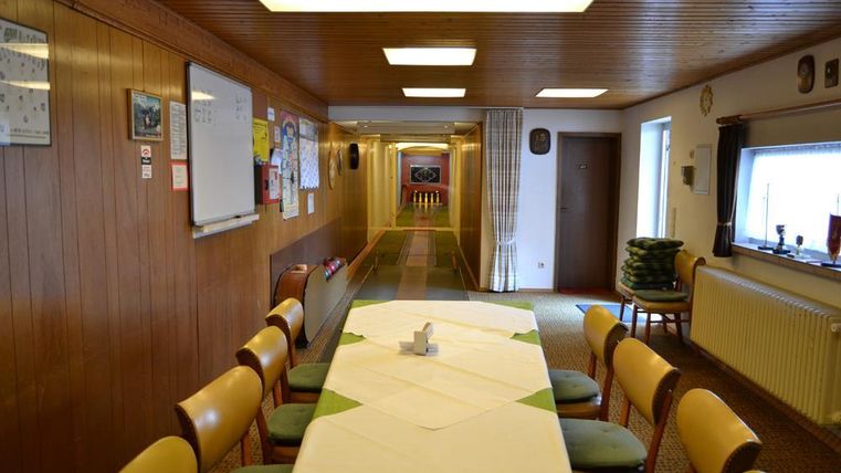 Een lange gang met houten wanden die naar een lichte kamer leidt. In het midden staat een gedekte tafel met stoelen.