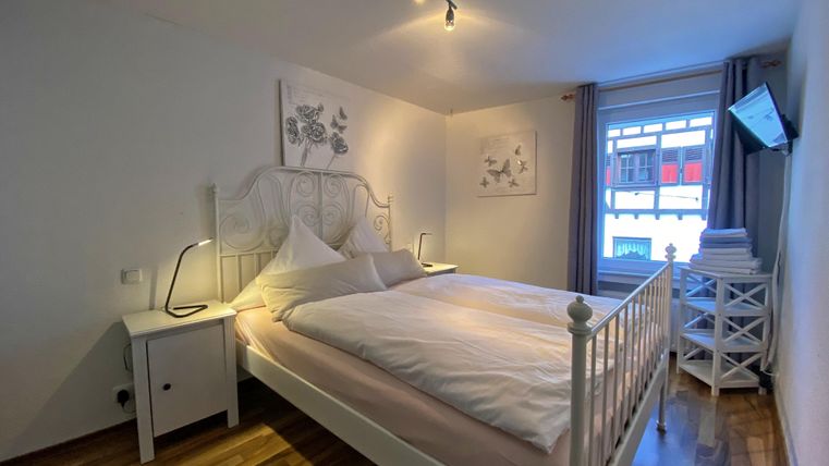 A cozy bedroom with a white bed and bright walls. The room has wooden flooring and a TV on the wall.