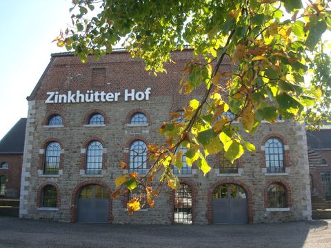 Een historisch gebouw genaamd Zinkhütter Hof met grote ramen en een stenen façade. Op de voorgrond zijn groene bladeren te zien.