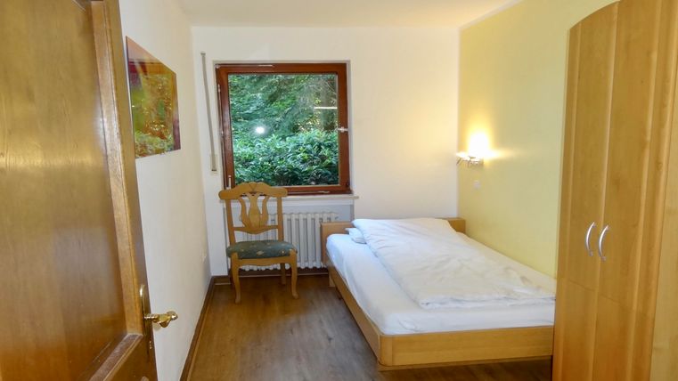 Ein einfaches Zimmer mit einem Einzelbett, einer Stuhl und einem Fenster, das auf eine grüne Landschaft blickt. Die Wände sind hell gestrichen und es gibt einen Holzschrank.