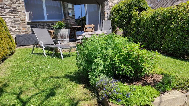 A garden with a beautiful seating area and umbrella. In the foreground, various plants and herbs are growing.