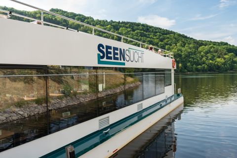 Een modern uitflugschip ligt rustig aan de oever van een meer. Op de achtergrond zijn groene heuvels en een blauwe lucht zichtbaar.