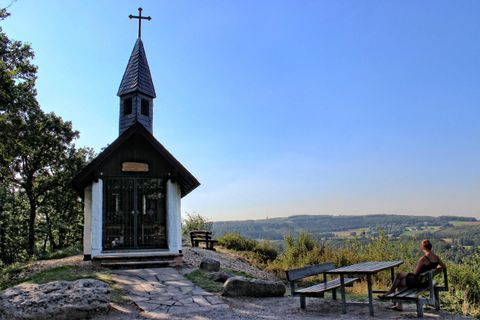 A small chapel with a cross stands on a hill. In the background, a picturesque landscape stretches under a clear blue sky.