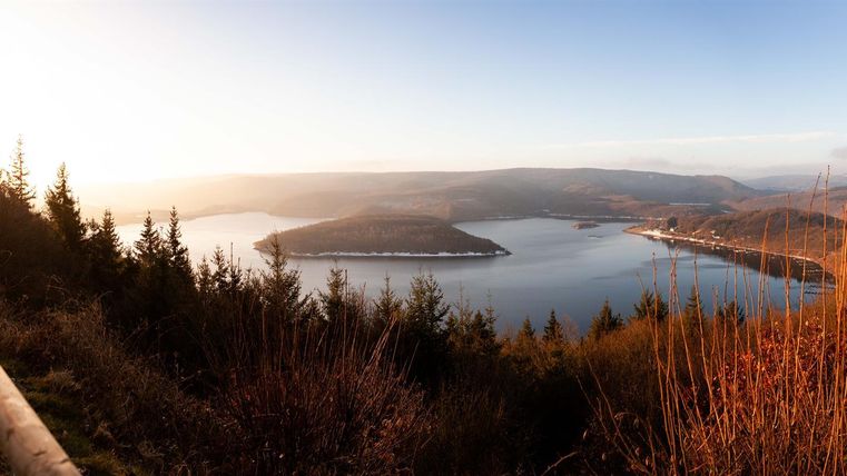 Een schilderachtig uitzicht op een meer, omringd door zachte heuvels en bossen. De lucht straalt in warme kleuren van de schemering.