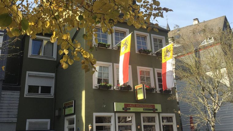 A green building with large windows and two flags. In the foreground, pedestrians and autumn leaves can be seen.