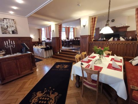 An elegant restaurant with wooden interior and laid tables. In the background, more tables and windows with curtains are visible.