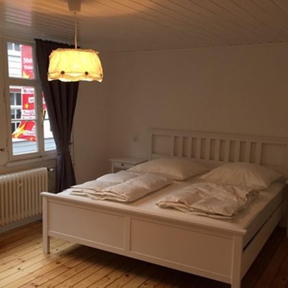 A bright bedroom with a white bed and two pillows. Large windows and a decorative light create an inviting atmosphere.