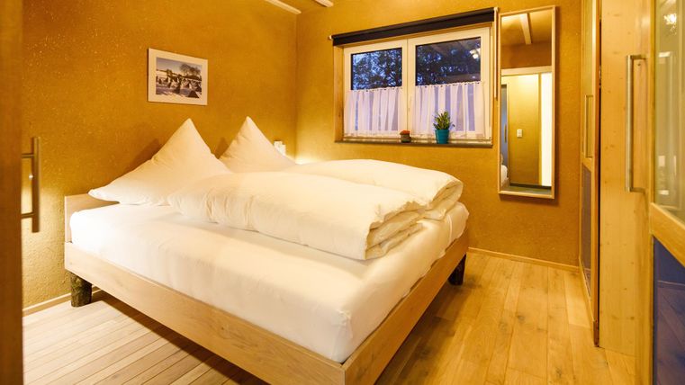 A cozy bedroom with a large bed and two white pillows. The walls are in warm earth tones, and a window lets in soft light.