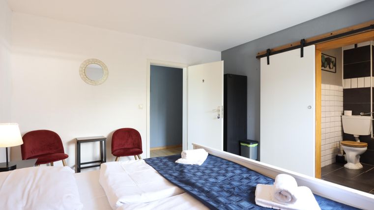A modern bedroom with a double bed and two red chairs. In the background, a bathroom and a large sliding door can be seen.