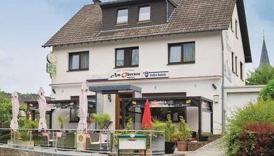 Ein gemütliches Gasthaus mit einer Terrasse, auf der Tische und Sonnenschirme stehen. Im Hintergrund ist ein ruhiger Ort mit grüner Landschaft zu sehen.