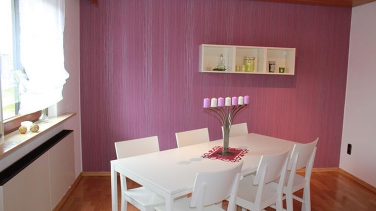 A modern dining area with a white table and seven chairs. The walls are purple and there is a windowsill with decorative elements.