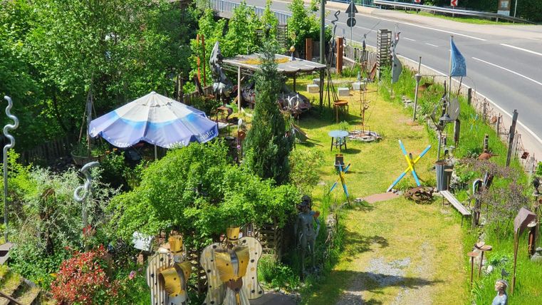Een originele tuin met verschillende sculpturen en een grote parasol. De groene omgeving en het pad leiden naar een straat.