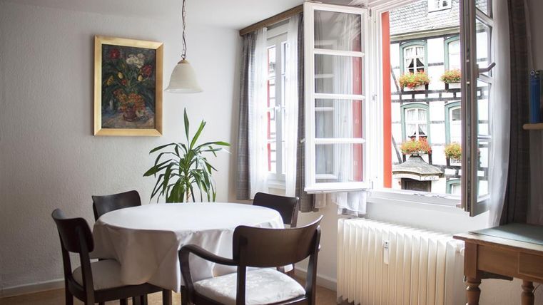 Een gezellige kamer met een ronde tafel en zwarte stoelen. Het raam is open en biedt uitzicht op kleurrijk versierde vensterbanken.
