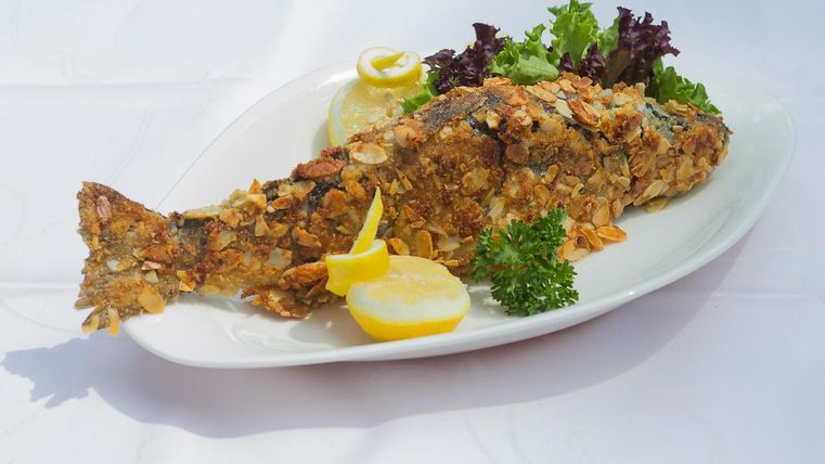 A fried fish on a white plate, garnished with lemon slices and fresh salad. The crispy crust is decorated with cornflakes.