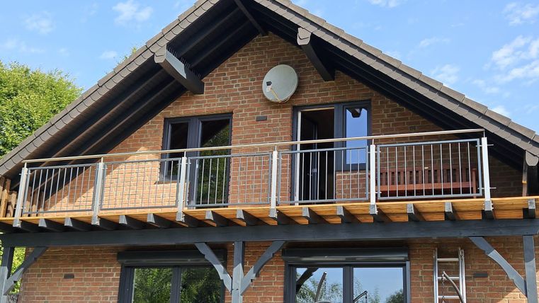 A modern house with a wooden porch and large windows. Visible railings are on the balcony and a satellite system is installed.
