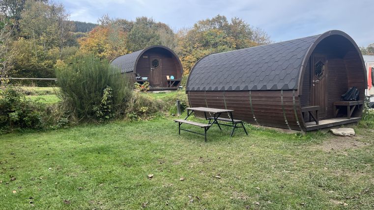 Twee gezellige hutten staan op een groene weide, omringd door bomen. Het weer is bewolkt, en er is een kleine zithoek buiten.