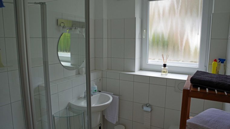 A modern bathroom with white tiles and a shower. The window provides daylight, and several personal care products are visible.