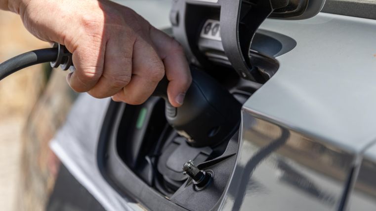 Een hand steekt een laadstekker in de oplader van een elektrische auto. Op de achtergrond zijn onscherpe natuurlijke elementen zichtbaar.