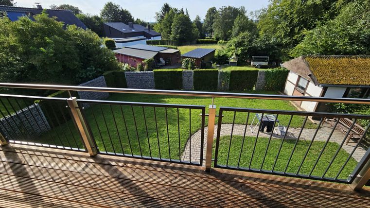 A view from a balcony of a beautiful garden with green grass and some trees. In the background, houses and a paved area can be seen.