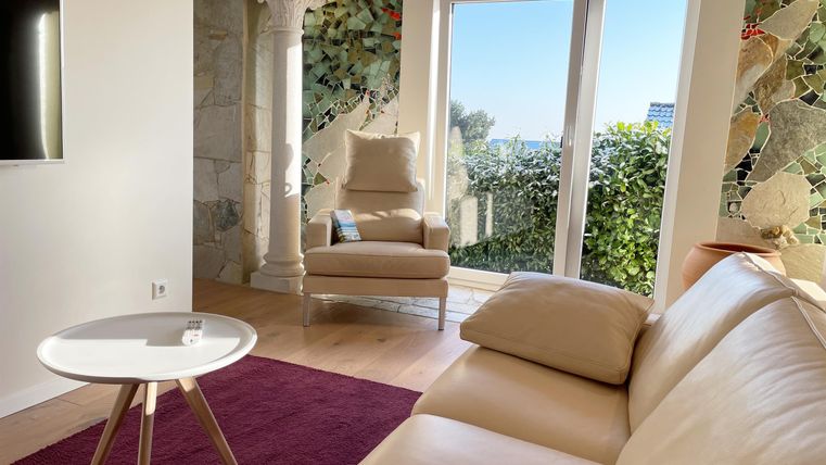 A modern living room with a beige sofa and a small white table. Large windows offer a view of the green surroundings and let in plenty of daylight.