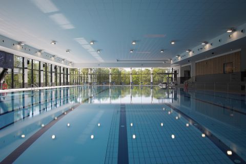 A modern indoor swimming pool with a clear water basin. Large windows let in plenty of daylight and offer a view of the outdoor area.