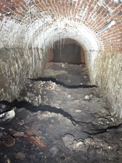 A dark tunnel with a vaulted brick roof. The floor is uneven and covered with trash and dirt.