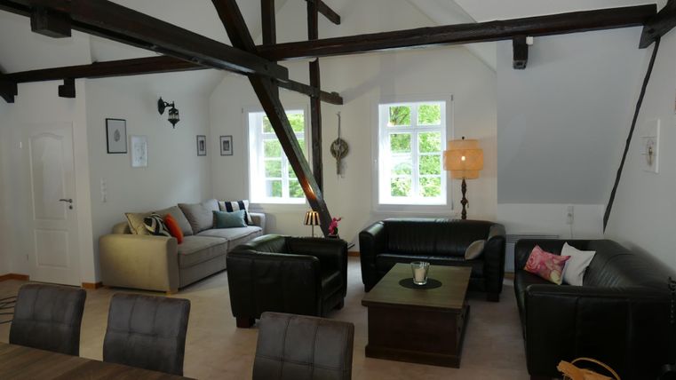 Een moderne woonkamer met hoge plafonds en zichtbare balken. Er zijn comfortabele banken, een tafel en grote ramen die veel licht binnenlaten.
