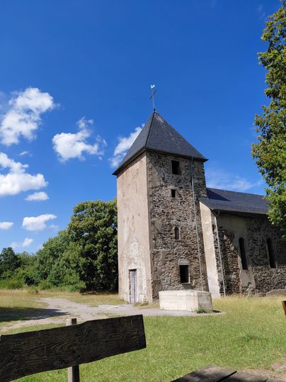 An old stone church with a pointed roof stands in nature. The sky is blue and there are some clouds.