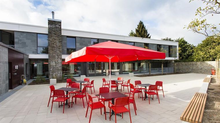 A modern terrace with red chairs and tables under a large red umbrella. In the background, there is an attractive building with large windows and a well-kept garden.