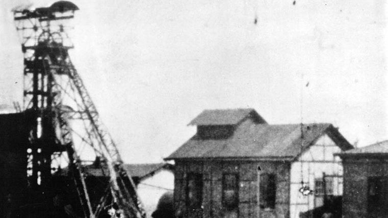 A historic industrial building with a headframe. The surroundings seem deserted and show signs of wear.