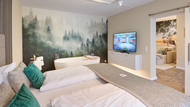 Ein modernes Schlafzimmer mit zwei Betten und einer gemütlichen Wandgestaltung in Waldoptik. Im Hintergrund befindet sich ein Fernseher und ein Badezimmer mit freistehender Badewanne.