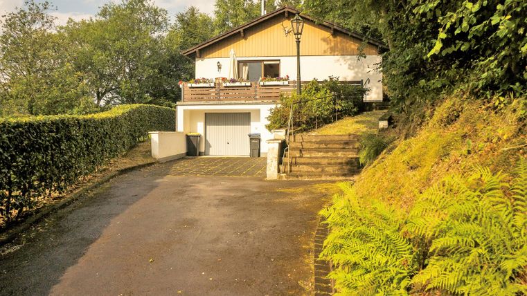 Ein gemütliches Haus auf einem leicht ansteigenden Grundstück, umgeben von Bäumen und Sträuchern. Der Weg zum Eingang ist gepflastert und von grünen Pflanzen gesäumt.