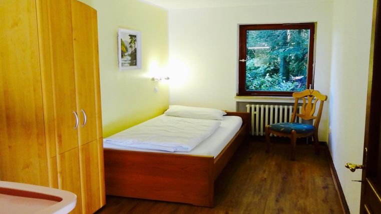 Ein einfaches Zimmer mit einem Bett, einem Schrank und einem Fenster, das auf einen grünen Garten blickt. Die Wände sind hellgelb, und es gibt einen Holzstuhl in der Ecke.