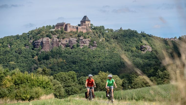 Twee fietsers rijden over een veldweg, terwijl op de achtergrond een indrukwekkend kasteel op een heuveltorent. Het landschap is groen en zonnig, ideaal voor outdooractiviteiten.