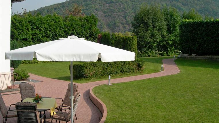 A well-maintained garden with a bright terrace and a large sunshade. In the background, green hedges and gentle hills can be seen.